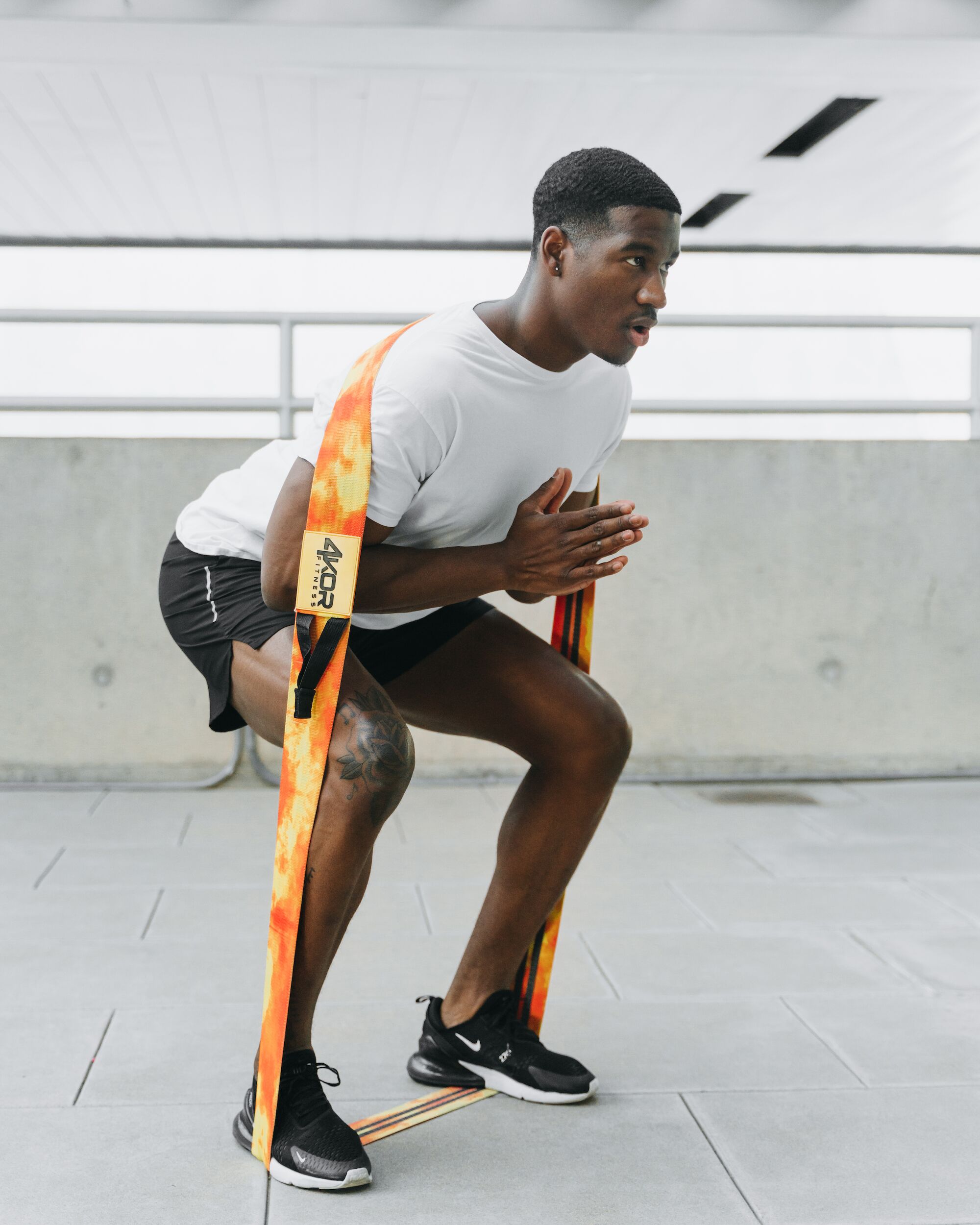 Male athlete performing resistance band squat with flame-print 4KOR Fitness flat resistance band anchored overhead outdoors for leg training