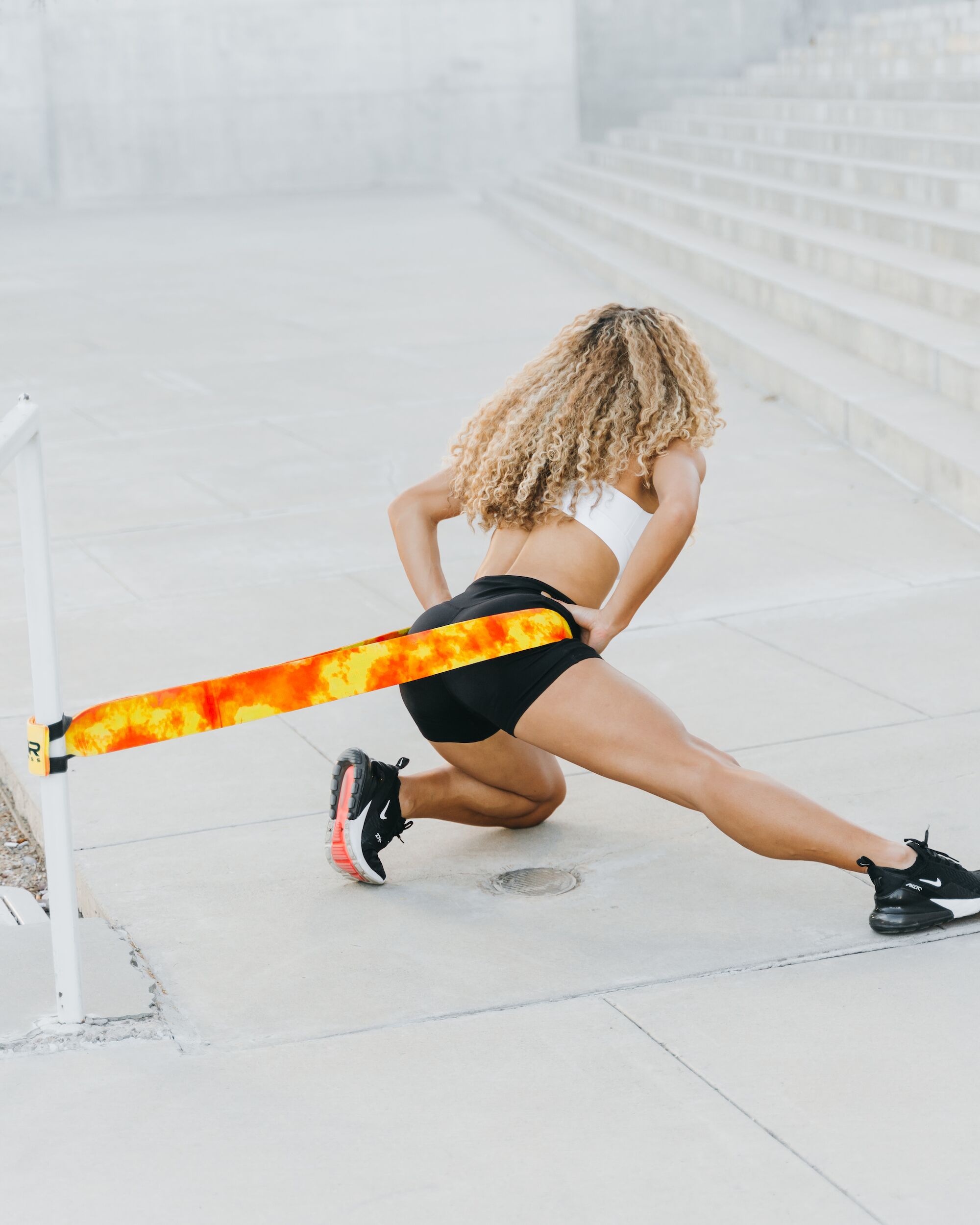 Female athlete performing lunge with flame-print 4KOR Fitness flat resistance band anchored to a post outdoors for hip flexor and glute training