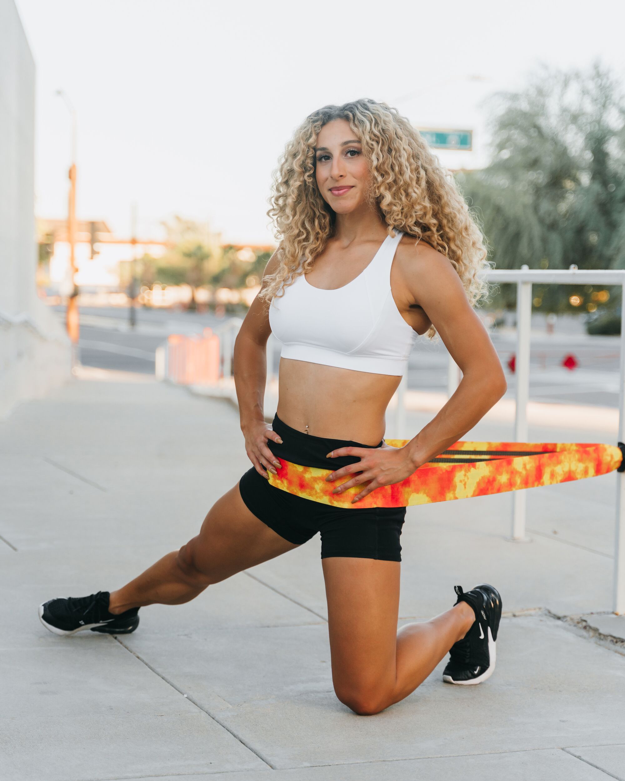 Female athlete performing lunge with flame-print 4KOR Fitness flat resistance band anchored to outdoor railing for hip flexor and glute training