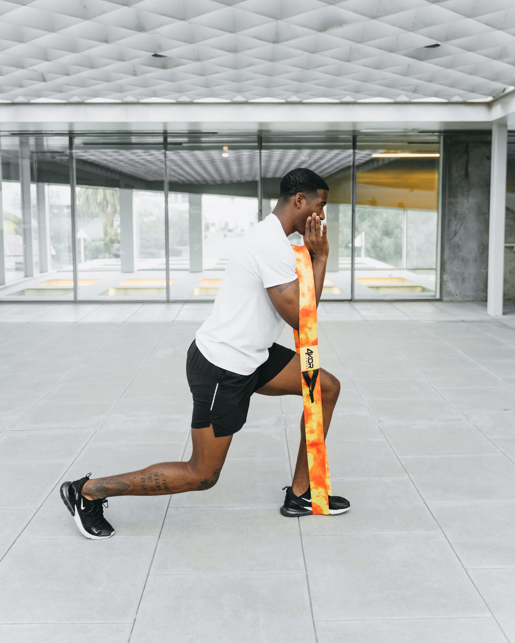 Male athlete performing lunge with flame-print 4KOR Fitness flat resistance band under foot outdoors for leg and glute strength training
