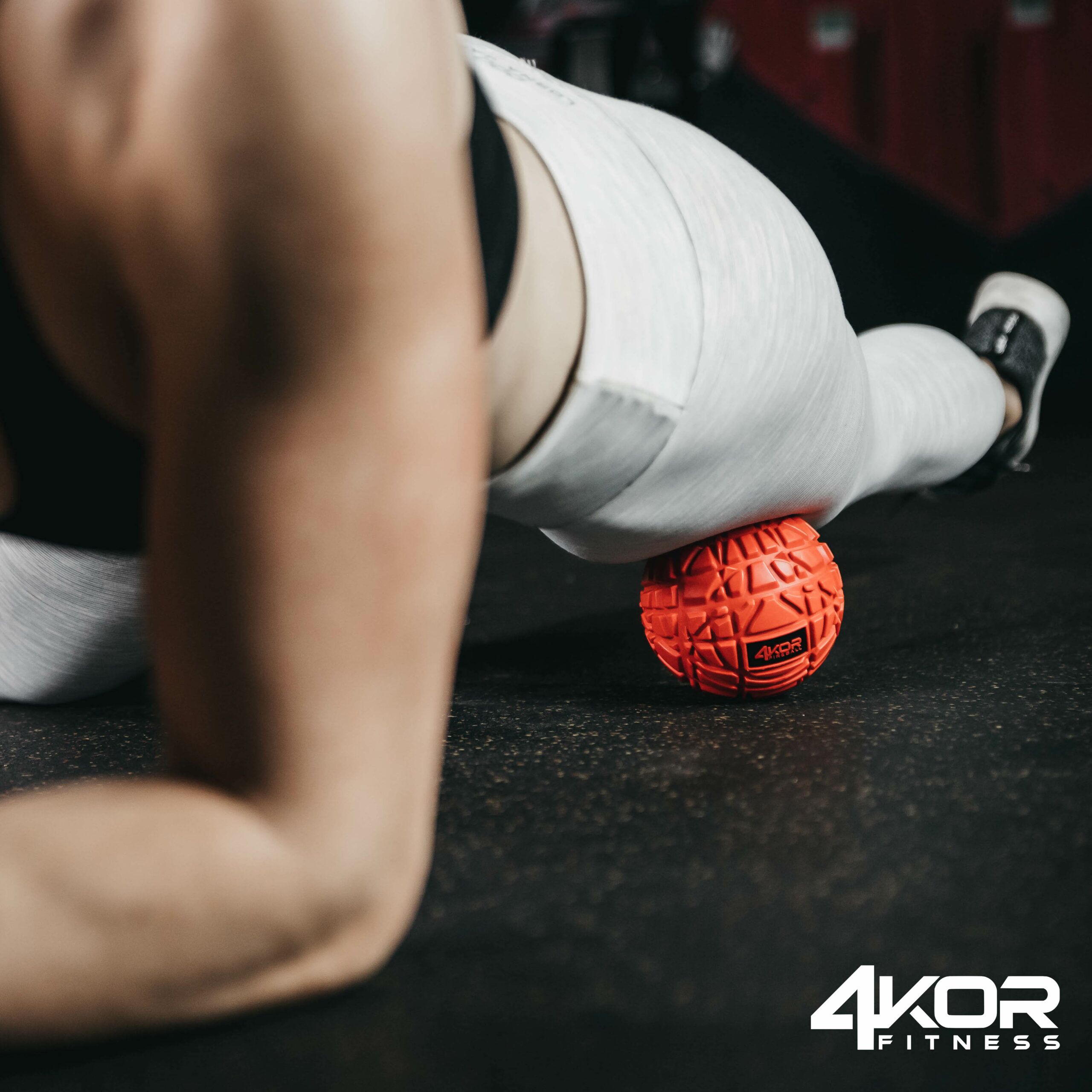Male athlete lying prone on gym floor pressing red 4KOR Fitness Fireball massage ball against shin for myofascial release, gym setting
