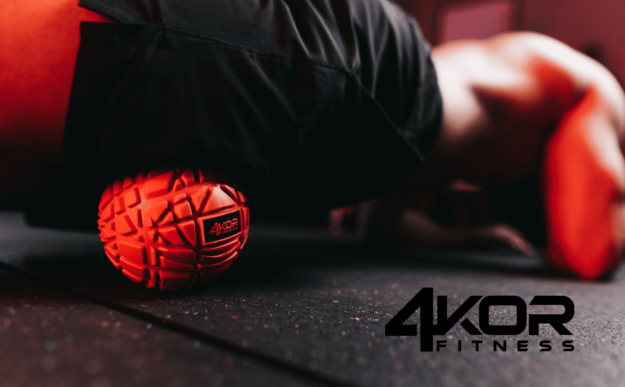 Close-up of red 4KOR Fireball massage ball with deep-grip texture on gym floor, blurred athlete in black hoodie in background, 4KOR Fitness logo