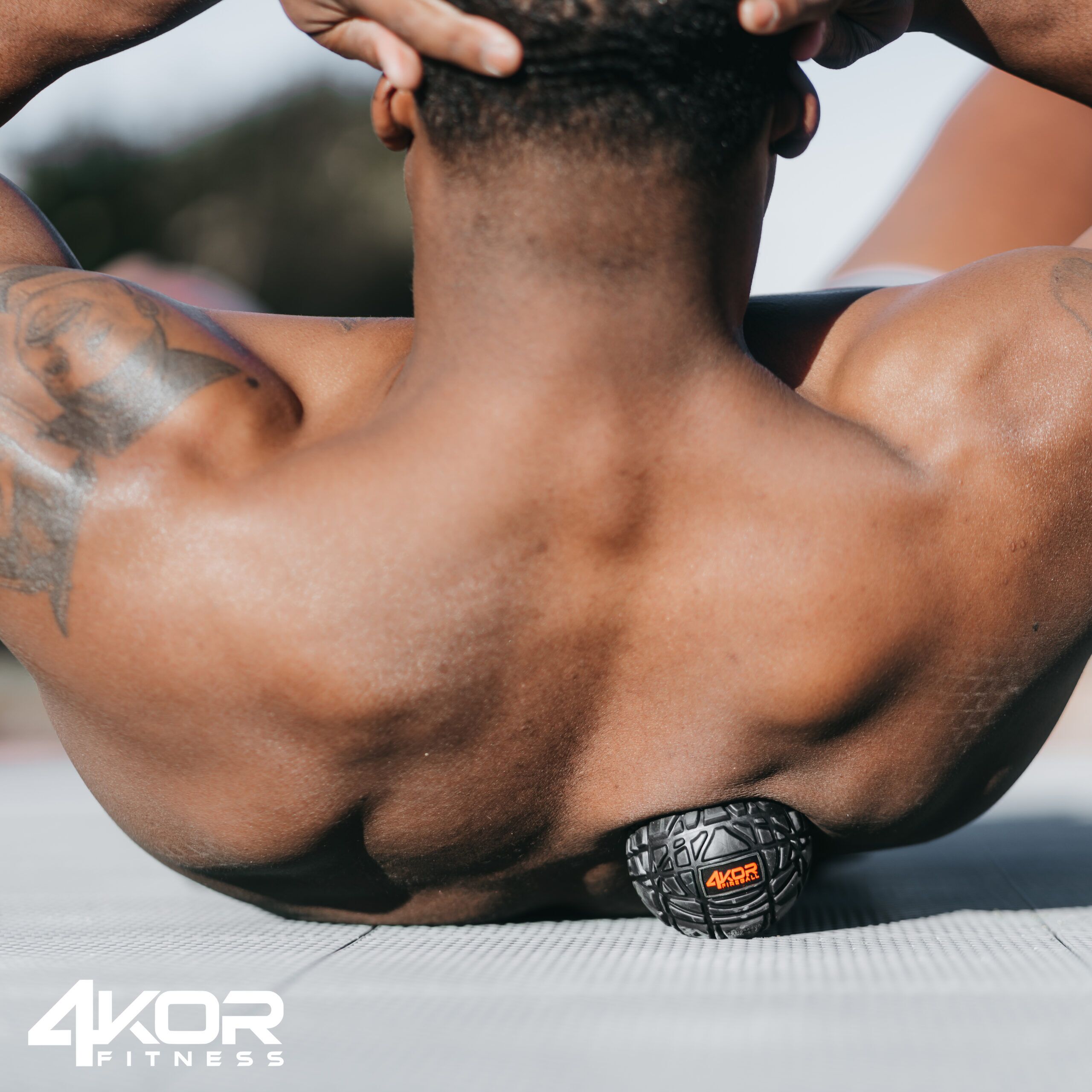 Shirtless male athlete lying on mat with 4KOR Fitness massage ball under lower back, hands behind head, rhomboid relief position