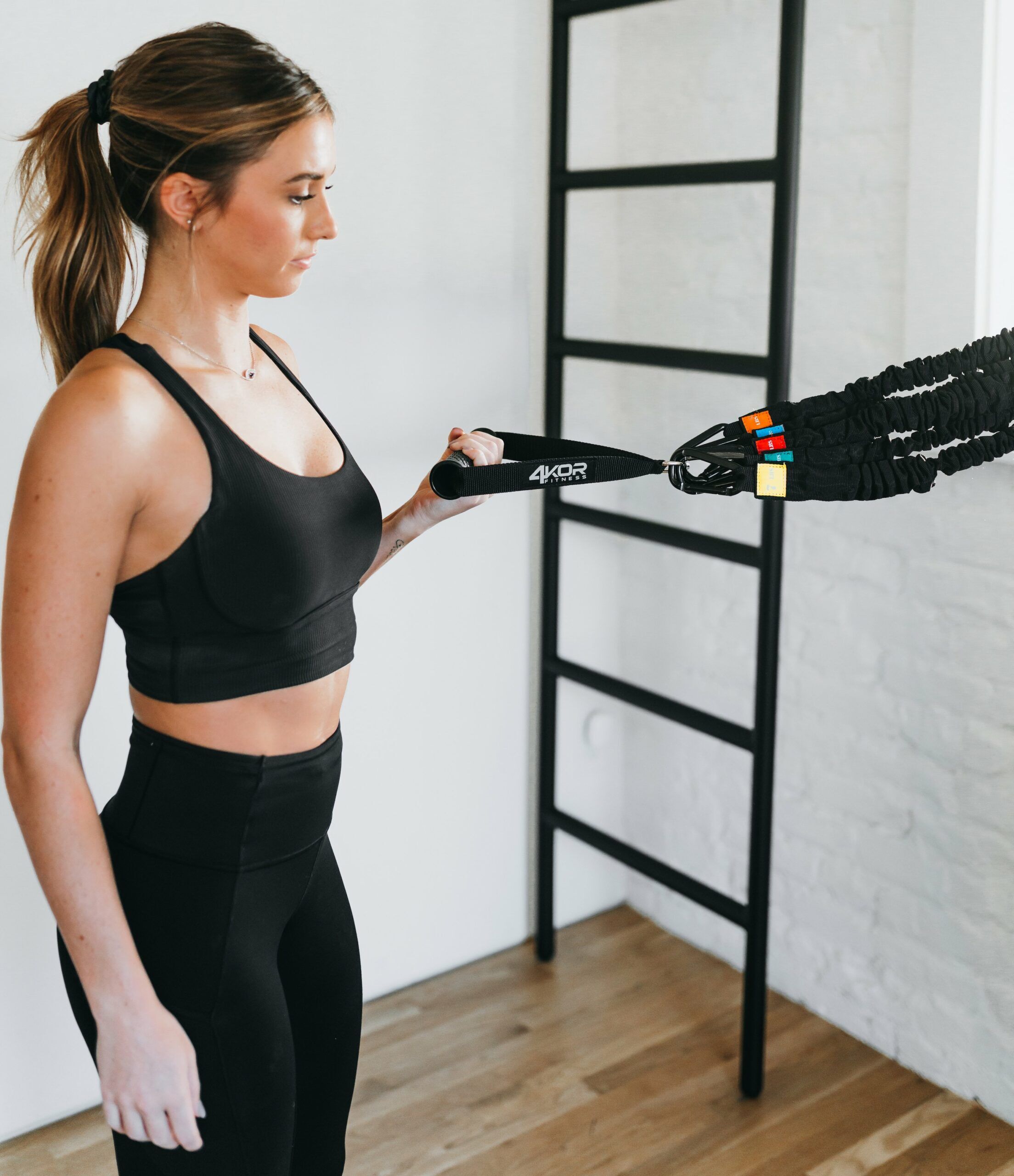 Female athlete in black sports bra gripping 4KOR Fitness exercise handle attached to bundled resistance bands, light studio with black ladder rack